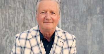 Glenn Tilbrook, an older man with grey slicked back hair, smiles as he's photographed wearing a navy shirt and a cream and blue plaid blazer