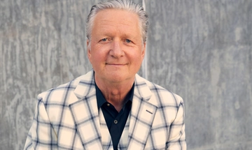 Glenn Tilbrook, an older man with grey slicked back hair, smiles as he's photographed wearing a navy shirt and a cream and blue plaid blazer