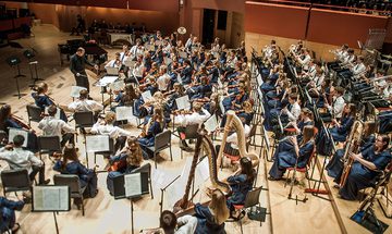 An orchestra of young musicians all in blue dress, or white shirts with blue ties, performing on The Anvil stage