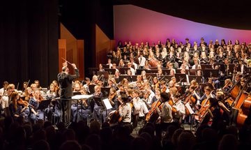 An orchestra of young musicians all in blue dress, or white shirts with blue ties, and a choir dressed in all black performing on stage