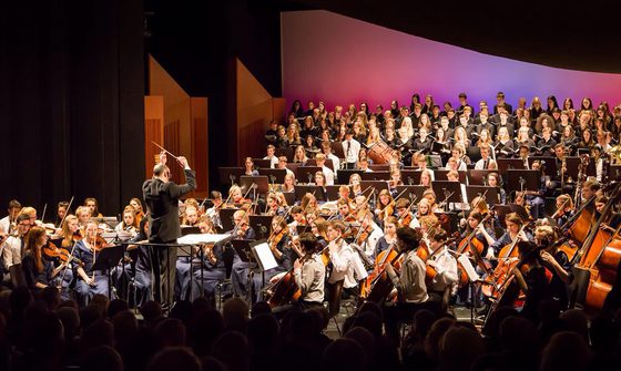 An orchestra of young musicians all in blue dress, or white shirts with blue ties, and a choir dressed in all black performing on stage