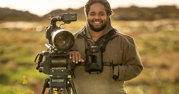 Hamza Yassin in a field at golden hour stood in a large coat with binoculars around his neck leaning on a large camera stood on a tripod