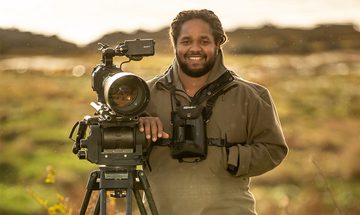 Hamza Yassin in a field at golden hour stood in a large coat with binoculars around his neck leaning on a large camera stood on a tripod