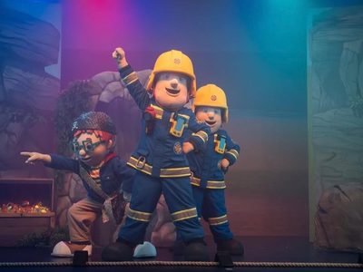 Fireman Sam pointing upwards on stage with two of his friends and a treasure chest in the background.
