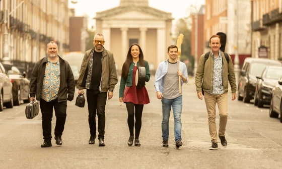 Five musicians, four men and one woman walk down the road in the the centre of a street. They all carry their instruments in cases and are dressed casually.