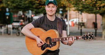 Jack Rutter, a white ginger man with short hair a short stubbly beard, in green cap and black t-shirt stood outside holding an acoustic guitar