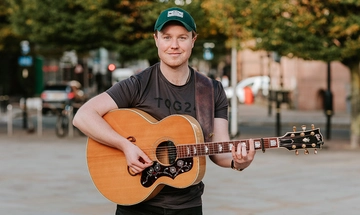 Jack Rutter, a white ginger man with short hair a short stubbly beard, in green cap and black t-shirt stood outside holding an acoustic guitar