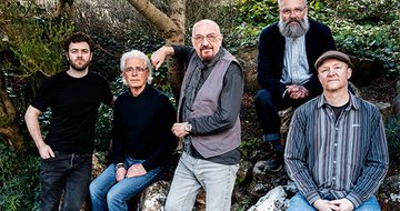 Five men all sit on a large rock in the woods. All are dressed in casual clothing in shades of grey, black and blue.