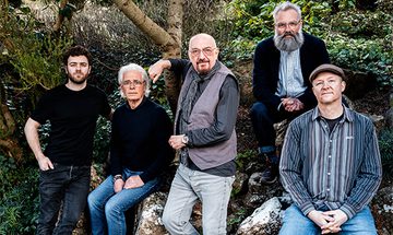 Five men all sit on a large rock in the woods. All are dressed in casual clothing in shades of grey, black and blue.