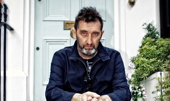 Jimmy Nail sat on a doorstep of a white house with a pale blue door. He has a neutral expression and is wearing denim coveralls with a t-shirt underneath with sunglasses hooked on them