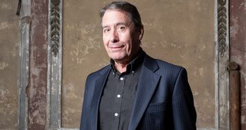 Jools Holland smiling softly in a dark suit jacket with a black shirt. He is stood in front of a weathered wall