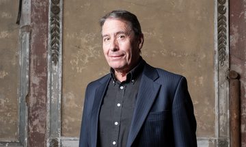 Jools Holland smiling softly in a dark suit jacket with a black shirt. He is stood in front of a weathered wall