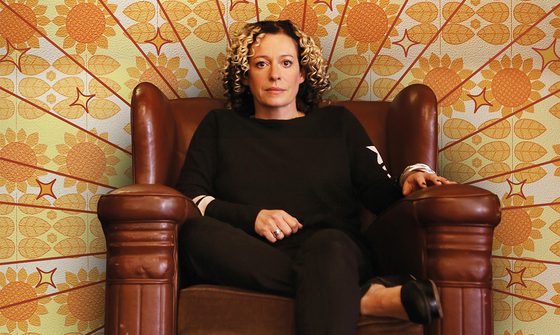 Kate Rusby in a large leather chair with 60s style wallpaper behind her