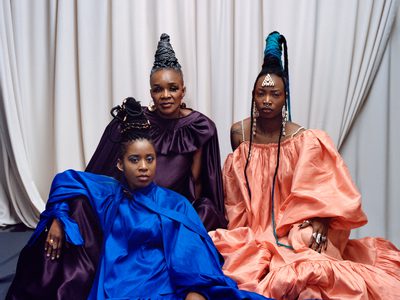 Three members of Les Amazones d'Afrique are sat in lounging positions on the floor staring at the camera with neutral expressions
