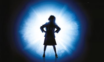 A silhouette of a young girl in a school uniform stood with her hands on her hips looking to the top left corner in front of a bright white and blue light