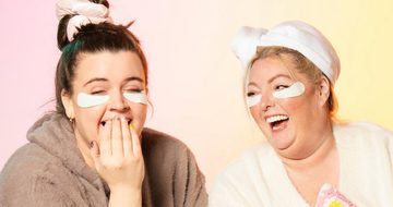 Tal Davies, a white woman with brown hair, and Mel Moon, a white woman with blonde hair, laughing together in dressing gowns with under eye masks on and their hair in a heatless curler and towel