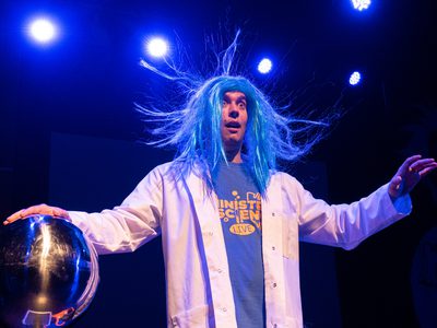 A male scientist in a lab coat wears a blue wig. He puts his hand on an electrified orb which makes the blue hair stand up on its end