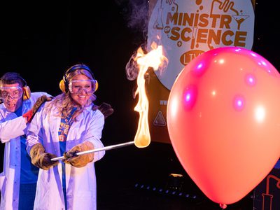 A male and female scientist, both in lab coats, the female scientist holds out a stick with fire on the end towards a large red balloon