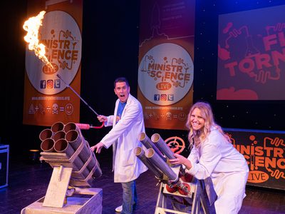 A male and female scientist in lab coats stand behind cannons, the man holds a fire lighter