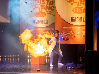 A scientist in a lab coat jumps back as flames explode on stage in front of them