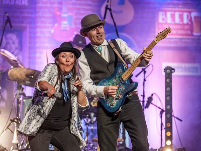 A man and woman performing together, the man is playing a guitar, the woman is pulling a silly face and point at the camera with a microphone in her hand