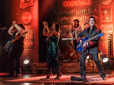 Four people on stage with microphones and instruments. They're all clapping apart from one man who is playing guitar
