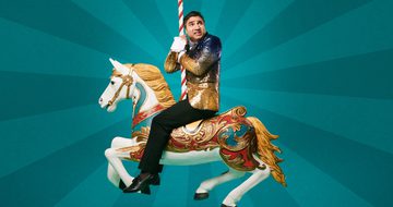 Nick Mohammed in a sequin gold and black blazer and black trousers sits on a merry-go-round horse, looking scared