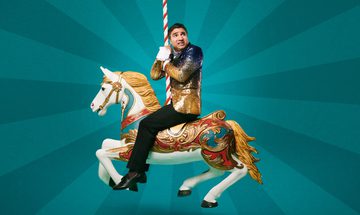 Nick Mohammed in a sequin gold and black blazer and black trousers sits on a merry-go-round horse, looking scared
