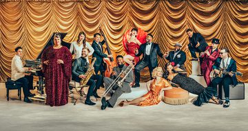 All 14 members of Postmodern Jukebox are all dressed in formal vintage evening wear and stood/sat on a concrete floor in front of a lavish golden curtain
