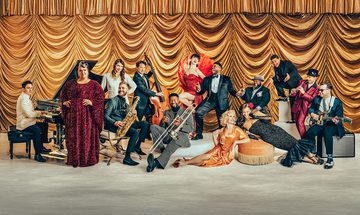 All 14 members of Postmodern Jukebox are all dressed in formal vintage evening wear and stood/sat on a concrete floor in front of a lavish golden curtain