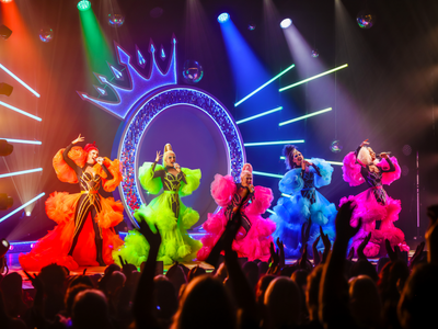 Five drag queens on a stage with a large glittery Q with a crown and multicoloured lights behind them. They're all singing in matching black body suits with coloured detailing as well as puffy skirts and sleeves. Each queen has a different colour being orange, green, pink, blue, and purple