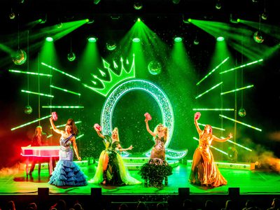 Four drag queens on a stage bathed in green light with a large glittery Q with a crown behind them. The queens are all singing or posing in ball gowns of different colours and also holding bubble guns. There is also a man sat at a piano with pink lights around playing and looking at the queens in awe
