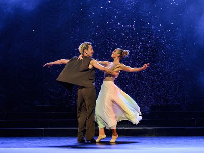 Carlos and Amy holding each other and spinning. Carlos is in a waistcoat and trousers and Amy is in a white and gold dress