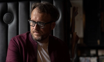 A dark photo of Robert Vincent, a white man with glasses and dark hair, sat down in a white t-shirt with a burgundy jacket