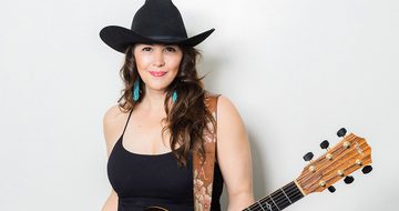 Sara Petite, a white woman with dark hair, wearing a black cowboy hat, black tank top, blue feather earrings, and holding an acoustic guitar strapped around her