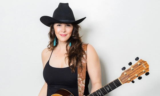 Sara Petite, a white woman with dark hair, wearing a black cowboy hat, black tank top, blue feather earrings, and holding an acoustic guitar strapped around her