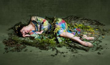 A woman in a green dress with splashes of other vibrant colours, lies on the floor surrounded by green leaves.