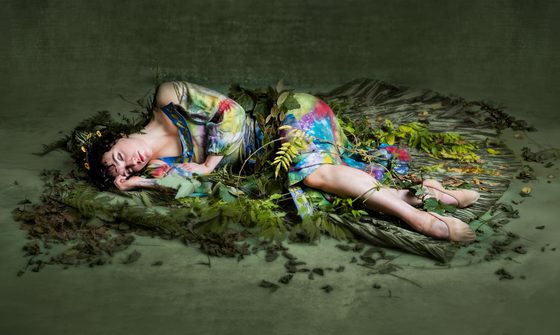 A woman in a green dress with splashes of other vibrant colours, lies on the floor surrounded by green leaves.