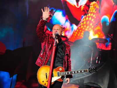 Steve Steinman in a red leopard-print jacket singing into a microphone, holding a guitar and raising one hand