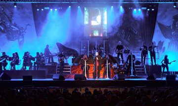 A rock band and orchestra on stage under blue lighting. Gothic artwork of dragons and a rider adorns the backdrop