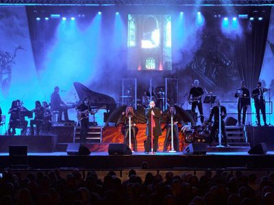 A rock band and orchestra on stage under blue lighting. Gothic artwork of dragons and a rider adorns the backdrop