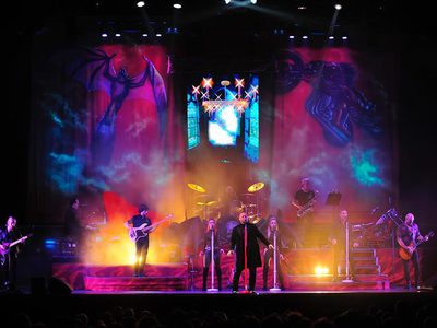 A rock band performing on stage with vibrant lighting, featuring a dragon and robot backdrop. Musicians play guitars and drums
