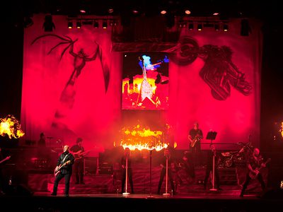 A rock band performing on stage with red and orange lighting, surrounded by flames