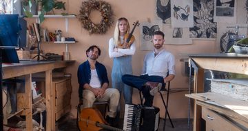 The three members of the band are gathered together, two sat down and the stood in the middle. They appear to be in an art studio or an office. It's full of drawings on the wall, lots of books and all the furniture is wooden.