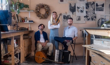 The three members of the band are gathered together, two sat down and the stood in the middle. They appear to be in an art studio or an office. It's full of drawings on the wall, lots of books and all the furniture is wooden.