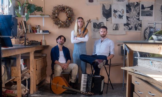 The three members of the band are gathered together, two sat down and the stood in the middle. They appear to be in an art studio or an office. It's full of drawings on the wall, lots of books and all the furniture is wooden.
