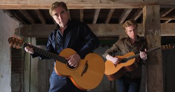 Brendan and Declan Murphy stand in an old building with exposed wooden bream as they play their acoustic guitars