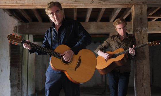 Brendan and Declan Murphy stand in an old building with exposed wooden bream as they play their acoustic guitars