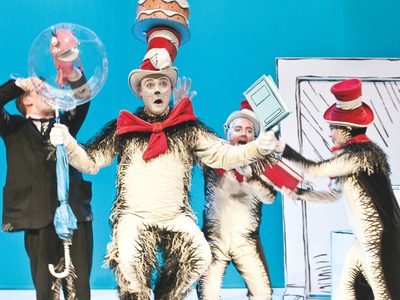 The fish, the Cat in the Hat and Thing 1 & 2 cause chaos inside Sally's house. The cat appears to be balancing a cake on top of his hat and Thing 1 & 2 are passing him books to hold.