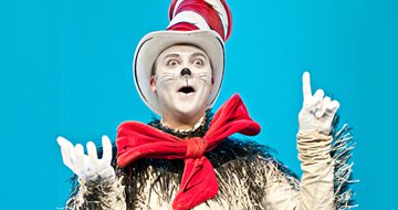 The Cat in the Hat, with his trademark red bow and red and white striped hat, points a finger to the sky like he has an idea.